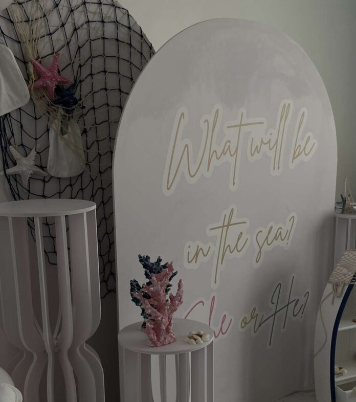 A photo shows the decorations of a gender reveal party with a sea theme. There is a table with one blue and one pink coral statue and a board with text that reads, “What will be in the sea? She or he?”